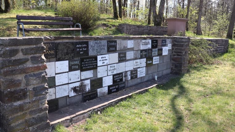 Ein Denkmal mit vielen Platten, die Namen und Texte tragen, um Erinnerung zu bewahren. Im Hintergrund sind Bäume und eine Bank zu sehen.