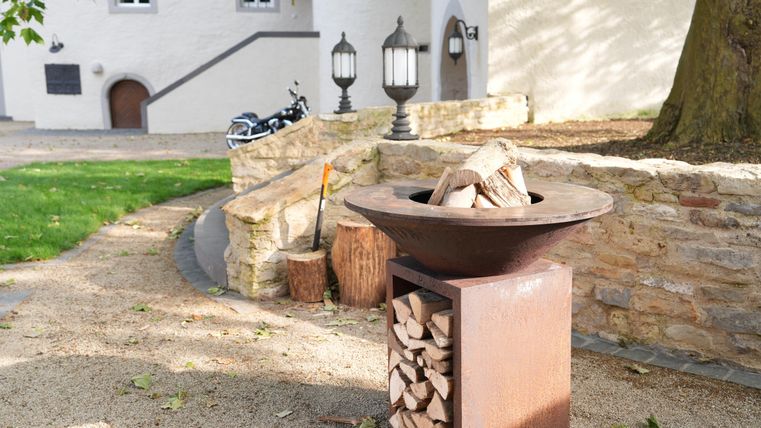 Ein Feuerstelle aus Metall mit Holz und einem rustikalen Design. Im Hintergrund sind eine Wand und einige Laternen zu sehen, die eine einladende Atmosphäre schaffen.