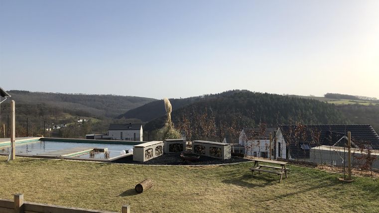 Ein ruhiger Ausblick über sanfte Hügel und das Dorf im Tal. Im Vordergrund befindet sich ein gepflegter Garten mit Bänken und Spielgeräten.
