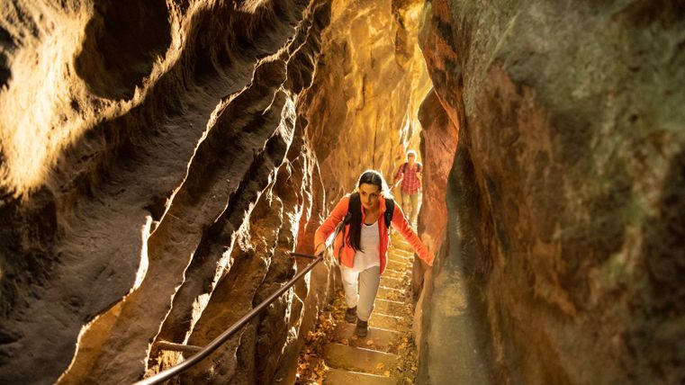 Zwei Personen durchqueren einen engen Felsdurchgang mit Treppenstufen und Handlauf.