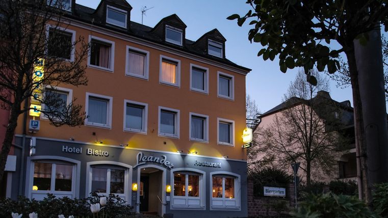 Un charmant bâtiment hôtelier avec une façade orange au crépuscule. L'entrée est accueillante, illuminée et entourée d'arbres et de fleurs.