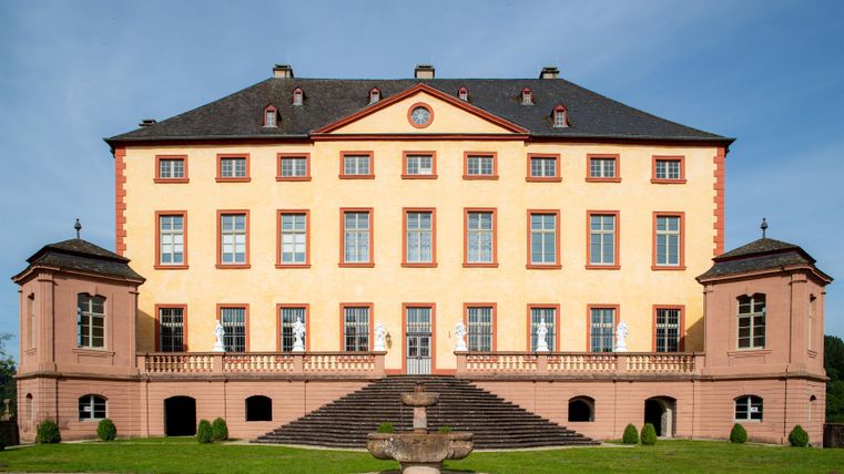 Ein historisches Schloss mit einer eleganten Fassade und großen Fenstern. Es liegt in einer grünen Umgebung mit einem gepflegten Garten.