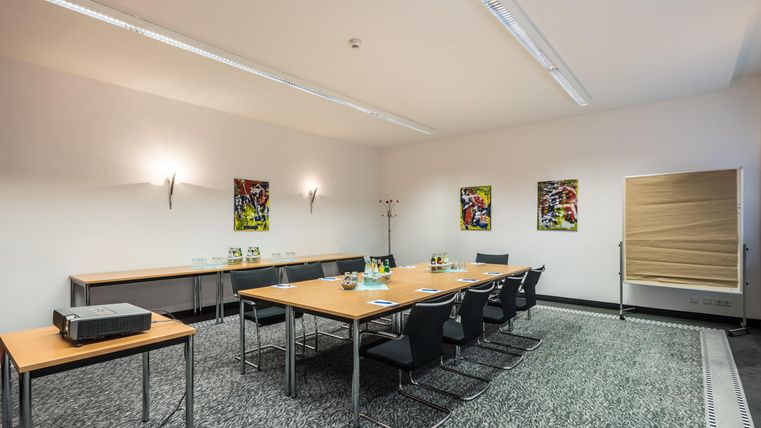 Een moderne vergaderruimte met een grote tafel en verschillende stoelen. Aan de muren hangen kleurrijke schilderijen, en er staan drinken op de tafel.