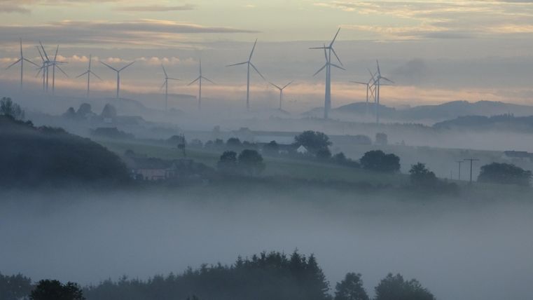  Een mistig landschap met windturbines op de achtergrond. De lucht is bewolkt en de nevel geeft de scène een mysterieuze sfeer.
