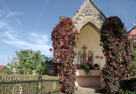 Eine kleine Kapelle in Wolsfeld, umgeben von roten Kletterpflanzen. Im Inneren sind religiöse Statuen zu sehen. Der Himmel ist klar und blau., © Tourist-Information Bitburger Land