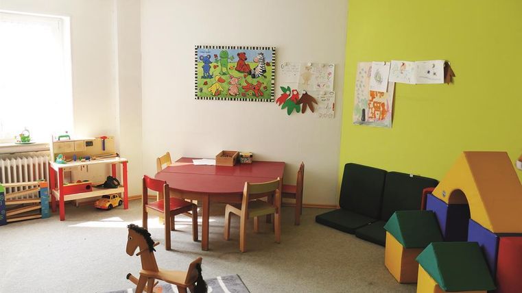 Ein bunter Spielraum mit einem roten Tisch, Stühlen und einem Spielhaus. Auf dem Boden steht ein Schaukelpferd und an der Wand hängen Kinderzeichnungen.