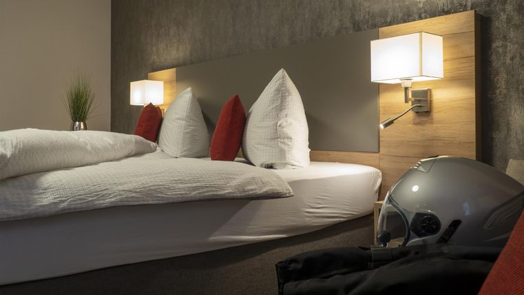 Ein gemütliches Schlafzimmer mit einem Doppelbett, modernen Nachttischlampen und einem Motorradhelm auf dem Boden. Die Wände sind in sanften Farben gehalten, was eine entspannende Atmosphäre schafft.