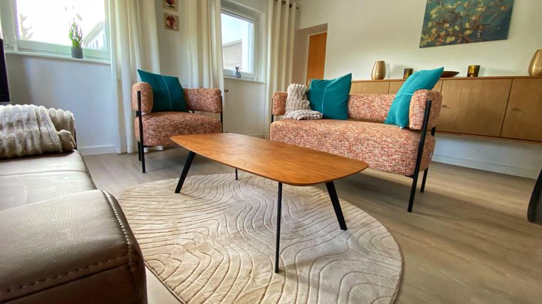 Ein gemütliches Wohnzimmer mit zwei bequemen Sesseln und einem niedrigen Holztisch. Die Wände sind hell gestrichen, und es gibt große Fenster mit Vorhängen.