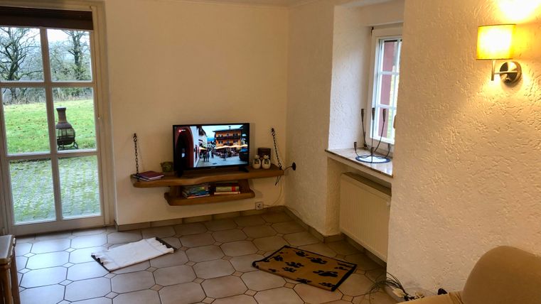 A cozy living space with a television on a shelf. Large windows allow plenty of daylight and offer a view of the greenery.