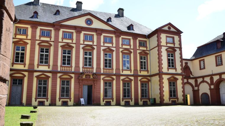Ein historisches Gebäude mit bunten Fassaden und großen Fenstern. Der Innenhof ist gepflastert und gut beleuchtet von der Sonne.
