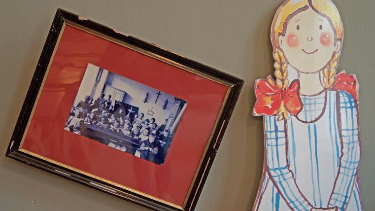 A framed photo of a group of children in a classroom hangs on the wall. Next to the picture is a painted figure of a girl with braids and a bow.