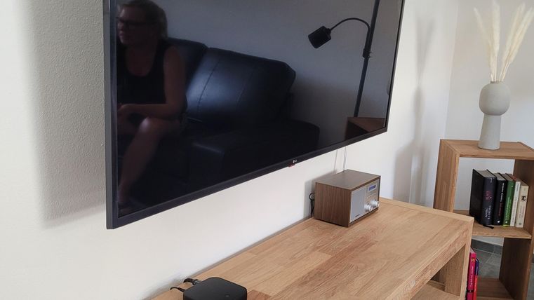 Ein moderner Fernseher hängt an der Wand über einem Holz-TV-Schrank. Auf dem Schrank befinden sich eine Fernbedienung und ein kleiner Lautsprecher.