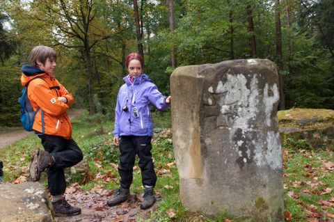 Zwei Kinder stehen im Wald neben einem alten Stein. Es ist Herbst und die Umgebung ist von bunten Blättern umgeben.