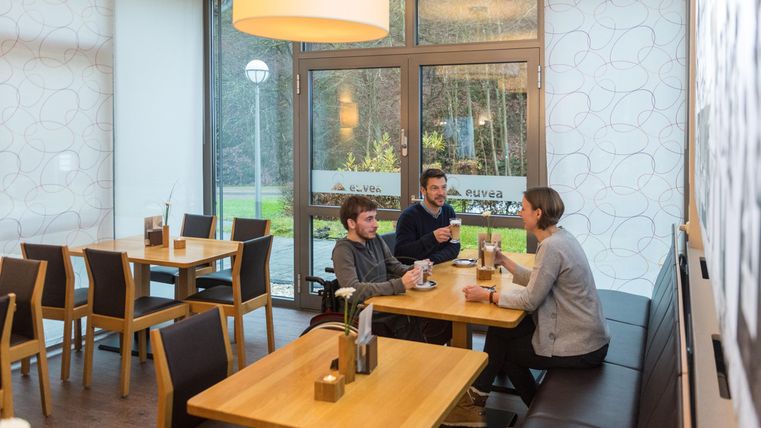 Een modern café met houten tafels en stoelen. Drie personen zitten samen en praten gezellig.