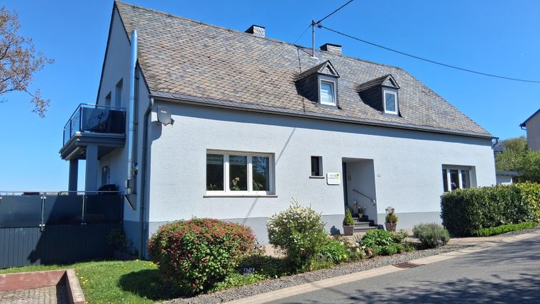 A bright, two-story house with a gray facade and a gable roof. The garden is well-maintained and there is a walkway in front of the house.