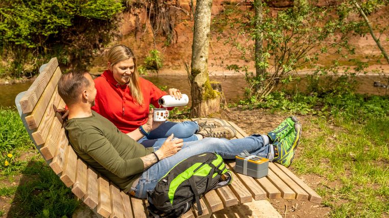 Ein Paar sitzt entspannt auf einer Bank im Freien. Sie genießen ein Picknick an einem malerischen Ort in der Natur.