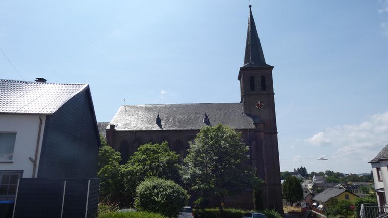 Eine alte Kirche mit einem hohen Turm steht hinter grünen Bäumen. Der Himmel ist klar und blau.