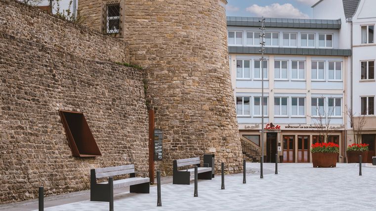 Ein historischer Turm aus Stein steht an einem Platz mit modernen Bänken. Im Hintergrund befinden sich mehrere Gebäude mit Fenstern und Grüntöpfen.