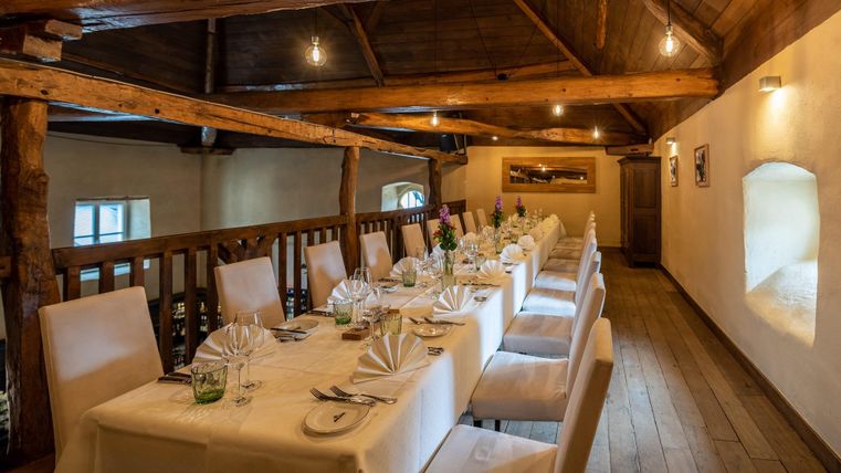 Een elegante eetruimte met lange tafels en witte tafellakens. Het houten balkenplafond en de zachte verlichting creëren een gezellige sfeer.