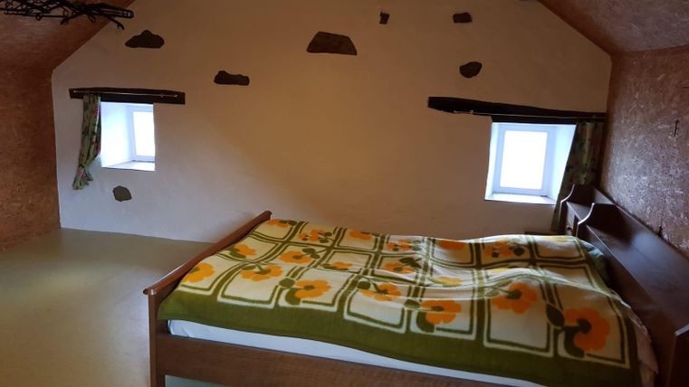 A cozy bedroom with a wooden bed and a colorful blanket. The walls are simply designed, with windows that let in light.