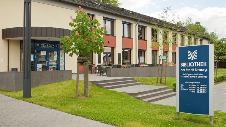 A modern library building with an inviting entrance. There are trees on the premises and an information board with opening hours.