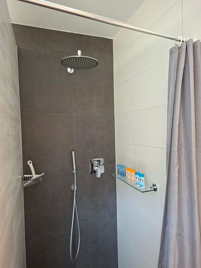 A modern shower with gray tiles and a large rain shower. Various care products are on a glass shelf.