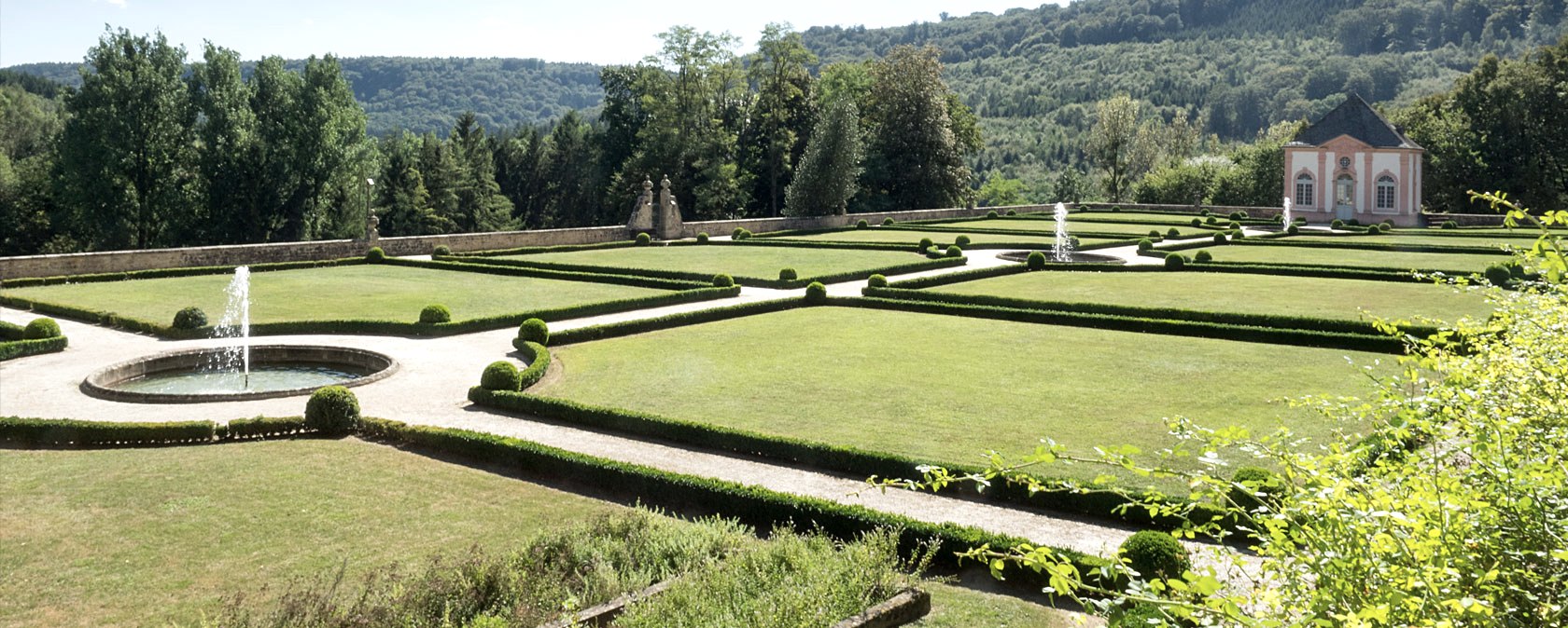 Ein gepflegter franz&ouml;sischer Garten mit symmetrischen Hecken, Springbrunnen und einem Pavillon im Hintergrund, umgeben von Wald., &copy; V. Teuschler