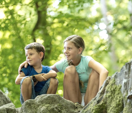 Zwei Kinder sitzen auf Felsen im Wald. Sie l&auml;cheln und schauen in die Ferne, umgeben von gr&uuml;nem Laub und nat&uuml;rlichem Licht., &copy; Felsenland S&uuml;deifel Tourismus GmbH