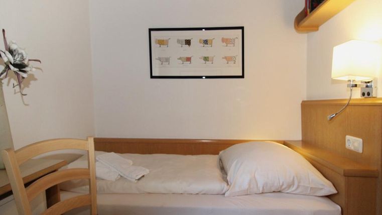 Ein einfaches Zimmer mit einem Einzelbett und einer gemütlichen Atmosphäre. An der Wand hängt ein gerahmtes Bild und eine Lampe sorgt für Licht.