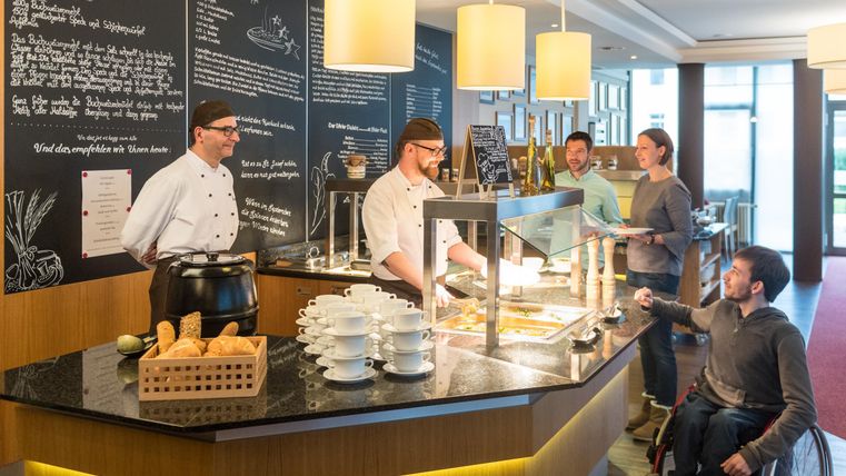Een moderne kantine met koks die gerechten uitserveren. Gasten worden vriendelijk bediend, waaronder ook een persoon in een rolstoel.