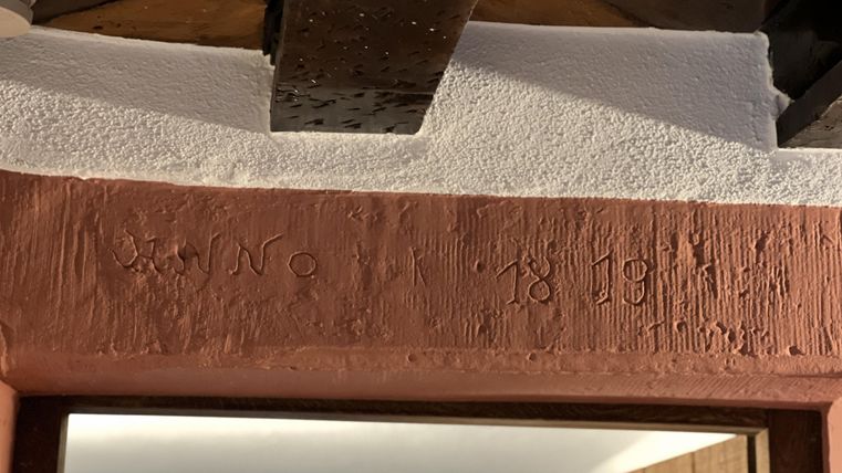 Een houten frame met een gegraveerd jaartal, dat vermoedelijk 1891 is. De muur heeft een rustieke, witte pleister.