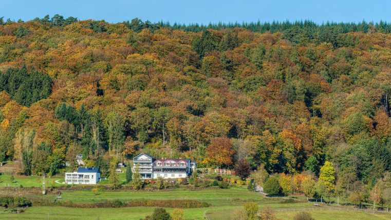 Ein malerisches Tal mit bunten Bäumen im Herbst. Im Vordergrund stehen zwei Gebäude, umgeben von der Natur.