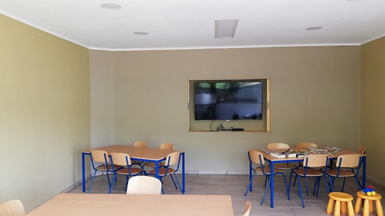 Een lichte kamer met grijze muren en een houten vloer. Er is een tafel met stoelen en een televisie aan de muur.