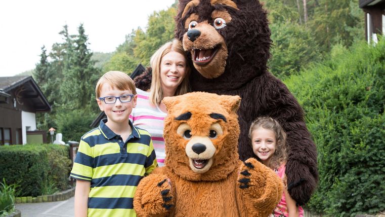 Zwei Bärenmaskottchen stehen mit vier Kindern in einem typischen Freizeitpark-Setting. Alle lachen und haben Spaß im Freien.