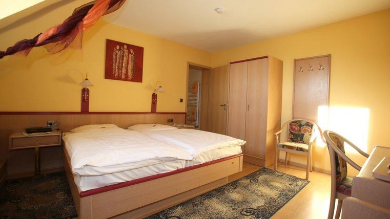 Ein gemütliches Zimmer mit einem Doppelbett und warmen gelben Wänden. Es gibt einen Schreibtisch, einen Stuhl und eine Garderobe.