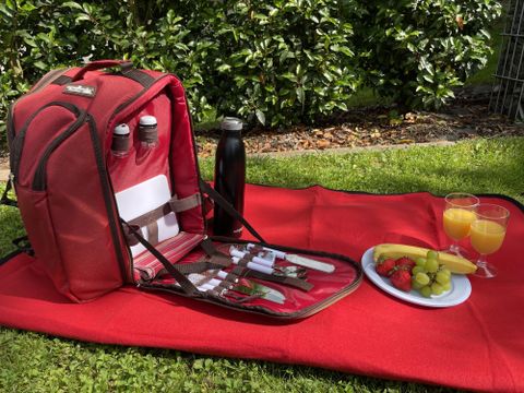 Un sac à dos de pique-nique rouge est posé sur une couverture dans la verdure. Il contient des couverts, des bouteilles et un espace avec des fruits frais et des boissons.