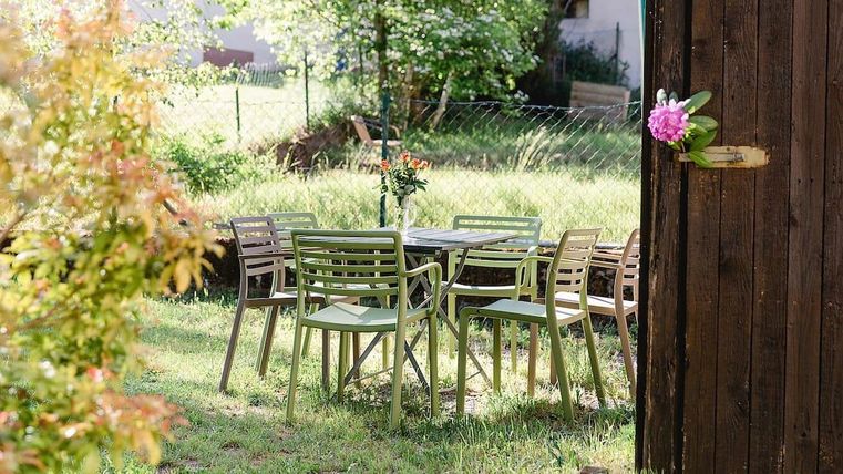 Ein gemütlicher Garten mit einem Tisch und Stühlen aus Holz. Im Hintergrund blühen Pflanzen und die Sonne scheint.