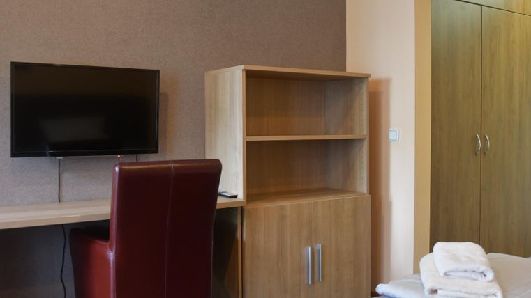 Een moderne slaapkamer met een bureau, een stoel en een televisie. Op de achtergrond staat een kledingkast en een bed met schone handdoeken.