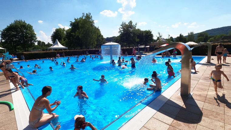 Ein belebter Swimmingpool mit vielen Menschen, die im Wasser spielen. Die Sonne scheint und es ist ein klarer blauer Himmel zu sehen.