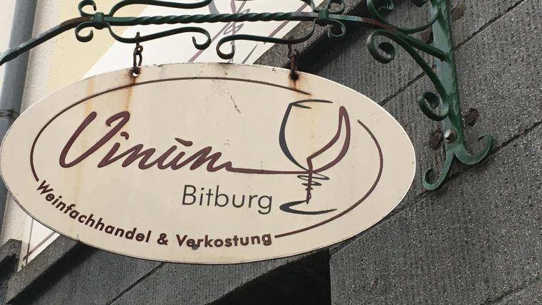 Een mooi bord met de tekst "Vinum Bitburg". Het toont een wijnspeciaalzaak en een proeverij.