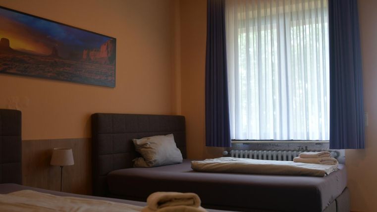 Een eenvoudige kamer met twee bedden en een raam dat veel daglicht binnenlaat. Aan de muur hangt een afbeelding van een landschap.