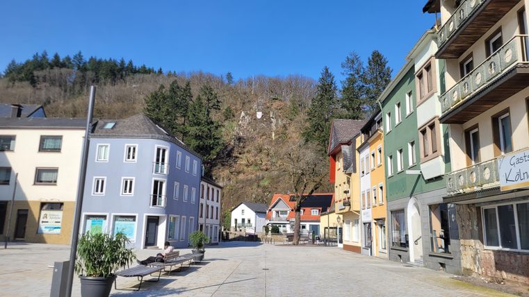 Ein ruhiger Platz mit bunten Gebäuden und einem klaren blauen Himmel. Im Hintergrund sind bewaldete Hügel sichtbar.