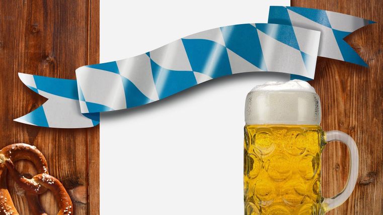 Ein Bierkrug mit Schaum und eine Brezel liegen auf einem Holztisch. Im Hintergrund ist eine weiße Fläche mit einem blauen und weißen Banner.