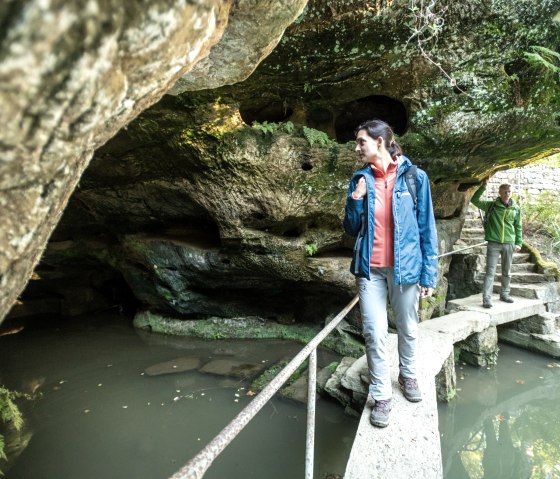 Zwei Personen auf einem schmalen Steg über einem Teich, umgeben von Felsen und Pflanzen. Eine Person trägt eine blaue Jacke, die andere eine grüne., © Eifel Tourismus, D. Ketz