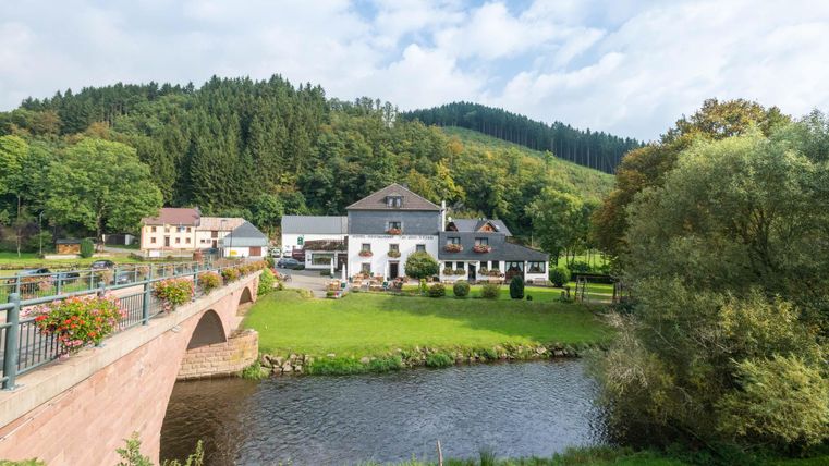 Un paysage pittoresque avec une rivière et un bâtiment historique. En arrière-plan, on peut voir des collines vertes et des forêts.