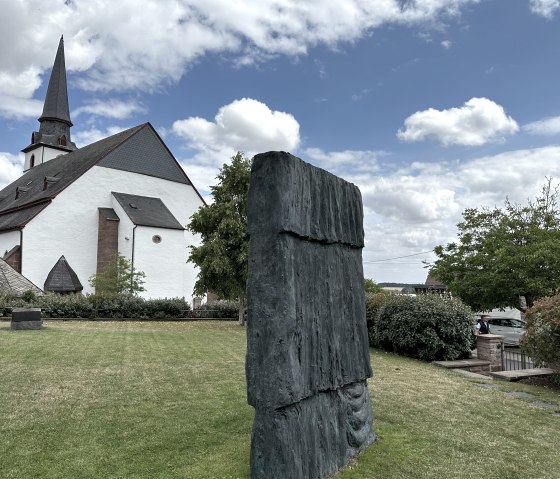 Eine Kirche mit spitzem Turm neben einer modernen Skulptur im Garten der Bibliothek G&uuml;nther F&ouml;rg in Weidingen., &copy; Felsenland S&uuml;deifel, Anna Carina Krebs