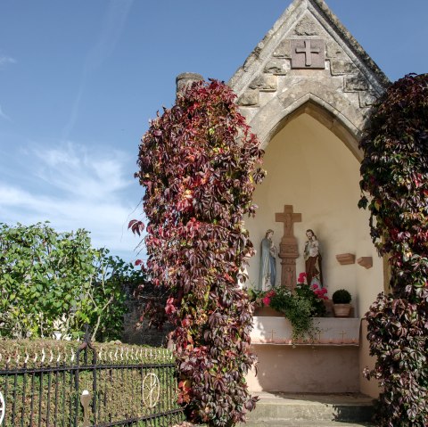 Eine kleine Kapelle in Wolsfeld, umgeben von roten Kletterpflanzen. Im Inneren sind religiöse Statuen zu sehen. Der Himmel ist klar und blau., © Tourist-Information Bitburger Land