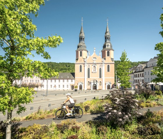 Ein Radfahrer f&auml;hrt vor der Basilika in Pr&uuml;m entlang, umgeben von gr&uuml;nen B&auml;umen und bunten Pflanzen, bei klarem Himmel., &copy; Eifel Tourismus GmbH, Dominik Ketz
