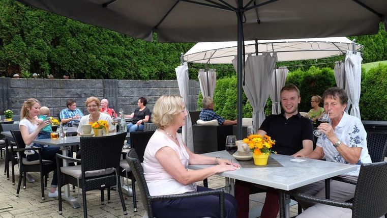 Eine gesellige Runde von Menschen sitzt in einem Außenbereich mit Tischen und Sonnenschirmen. Es gibt Blumen auf den Tischen und die Gäste genießen Getränke.