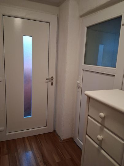 Une entrée lumineuse avec une porte blanche et une fenêtre. À droite se trouve une commode blanche sur un sol en bois.